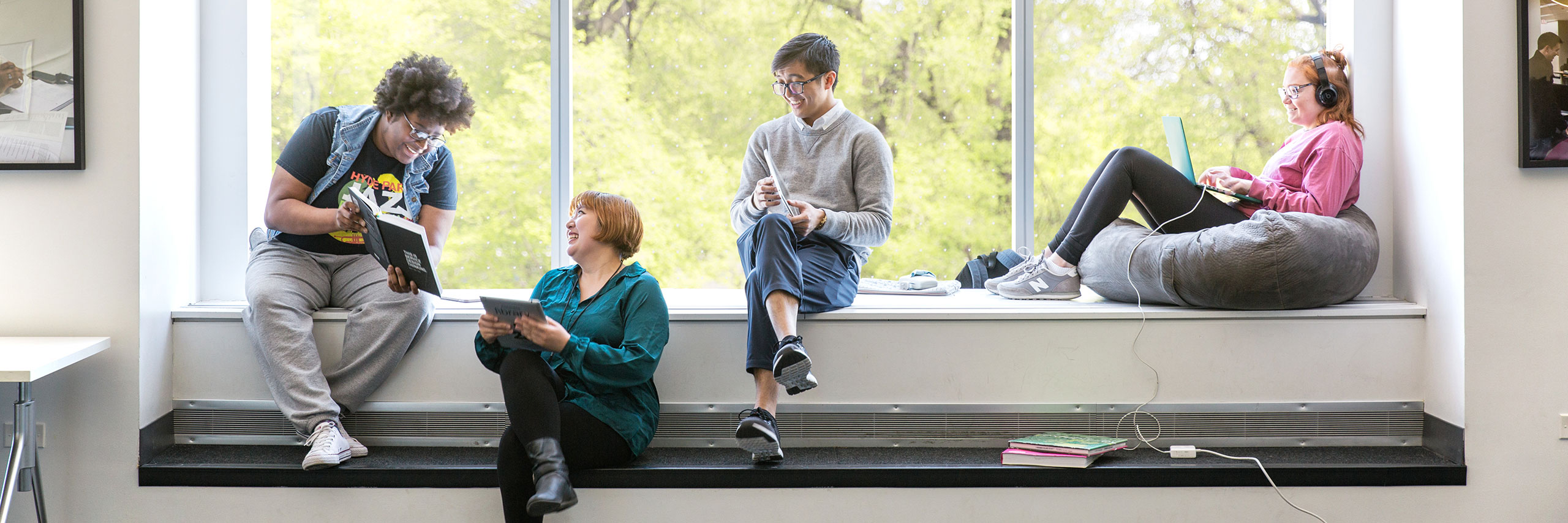 Four students sitting on a windowsill engaged in reading and using devices, with trees visible outside.
